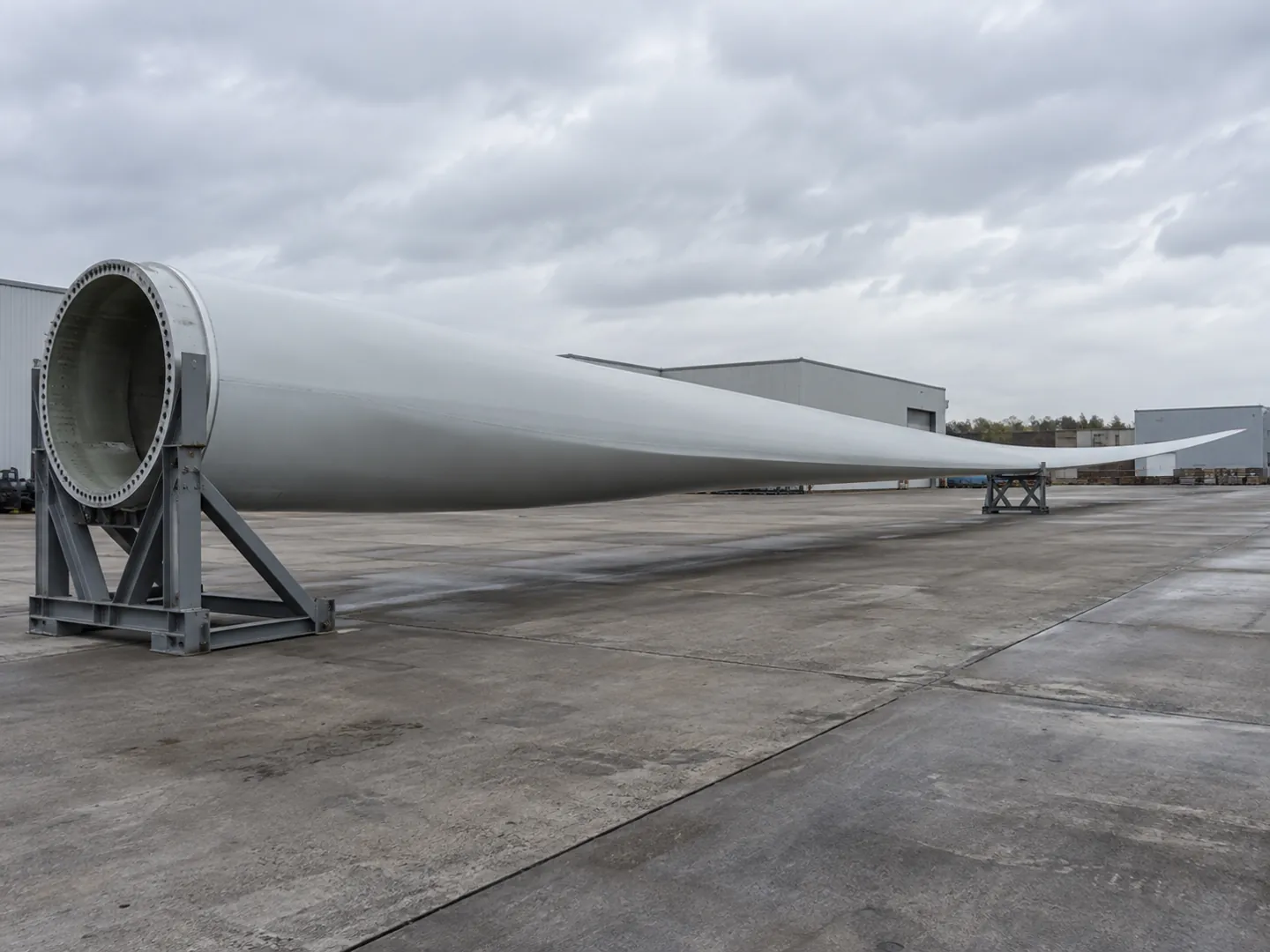A single long white wind turbine blade resting on supports in an industrial yard, its curved fibreglass shell catching the light